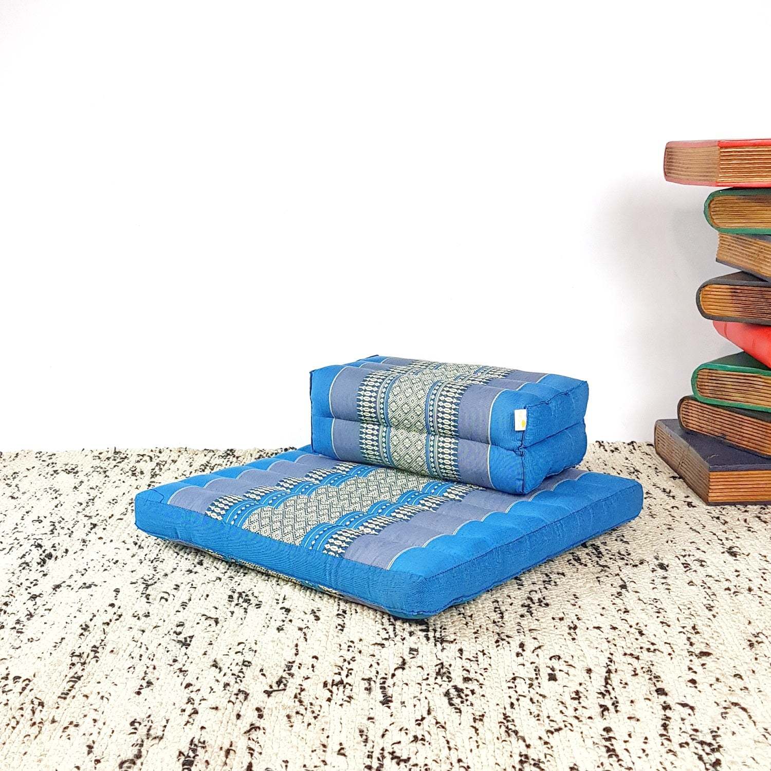 Blue Thai meditation cushion and seating block set with traditional patterns on a textured rug next to wooden stacked books