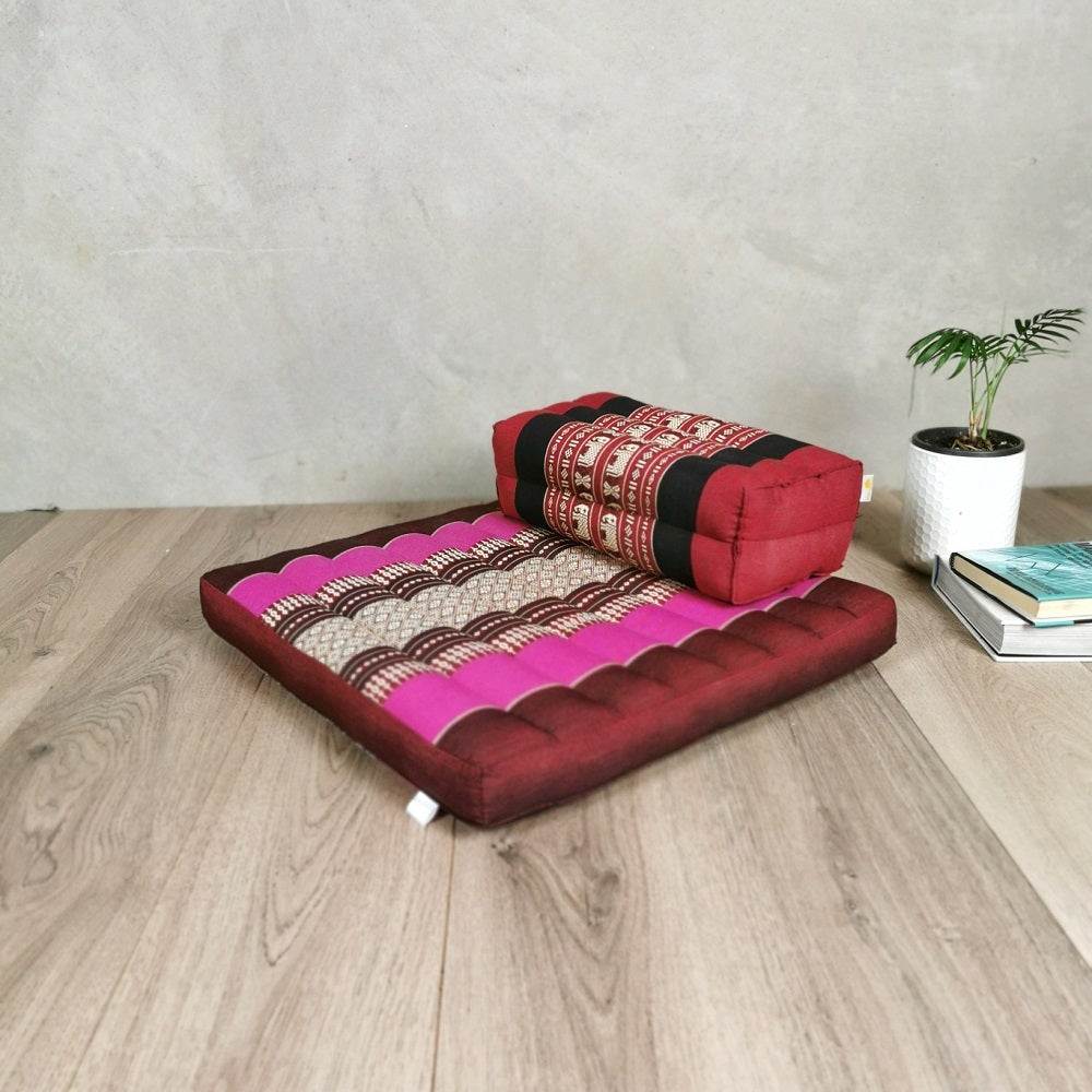Square red and pink Thai meditation cushion and seating block set on wooden floor with small plant and books nearby