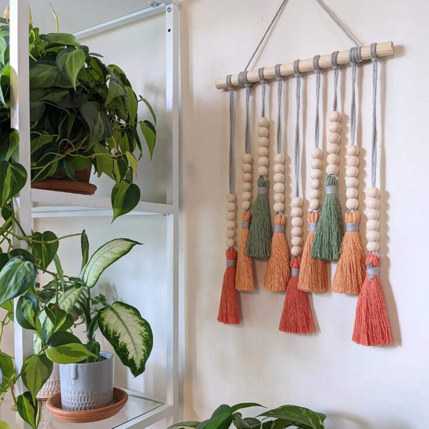 Colorful tassel wall hanging with wooden beads in a boho style displayed on a white wall beside green indoor plants