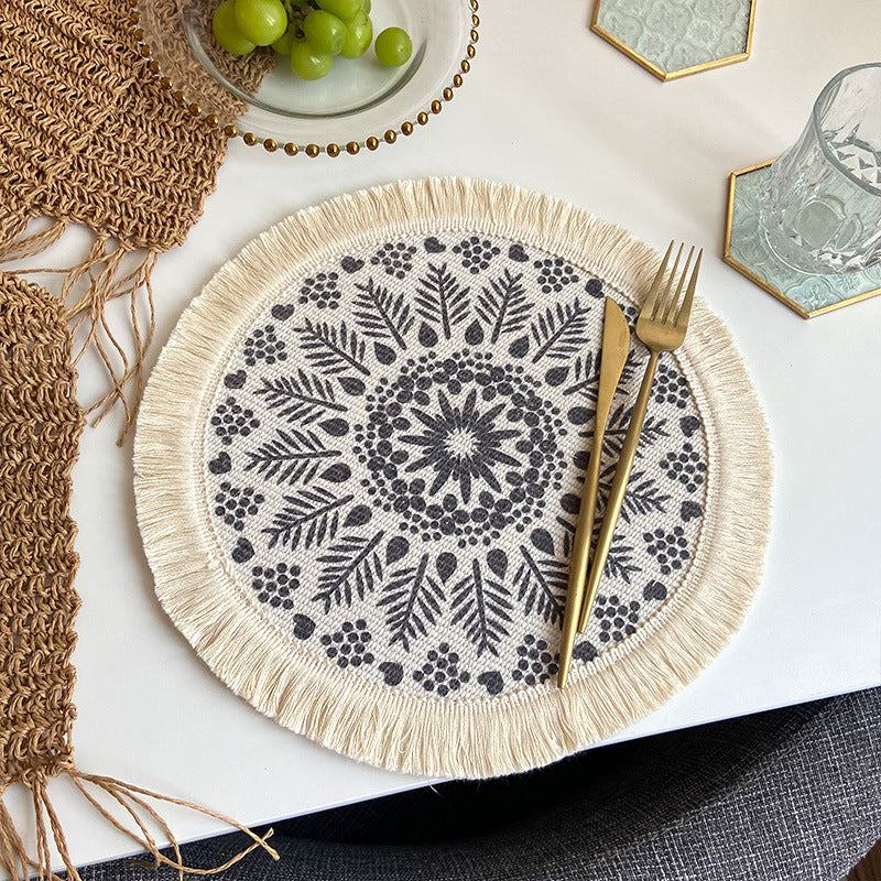 Beige and grey boho chic woven cotton placemat with mandala design and tassels on a white table with gold cutlery and decorative items.