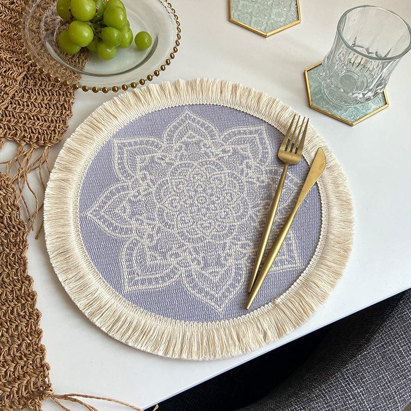 Round beige and purple boho chic woven cotton placemat with mandala design and tassels on white table with gold cutlery and glass.