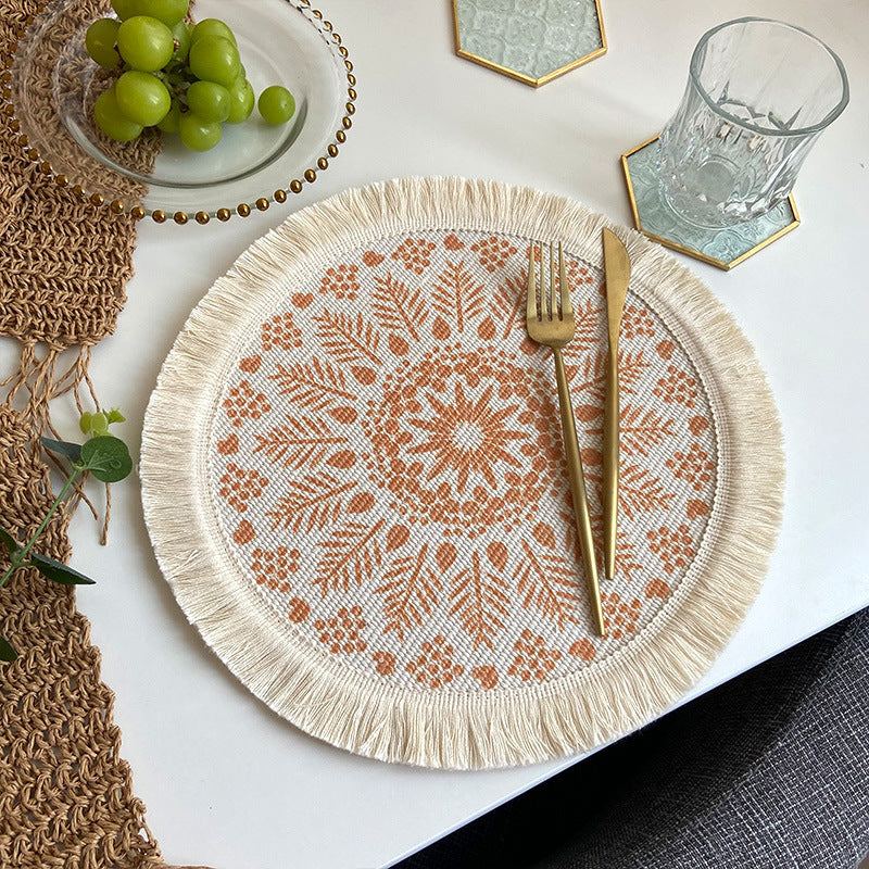 Beige and orange boho chic woven cotton placemat with mandala design and tassels on a white table setting with gold cutlery and glassware